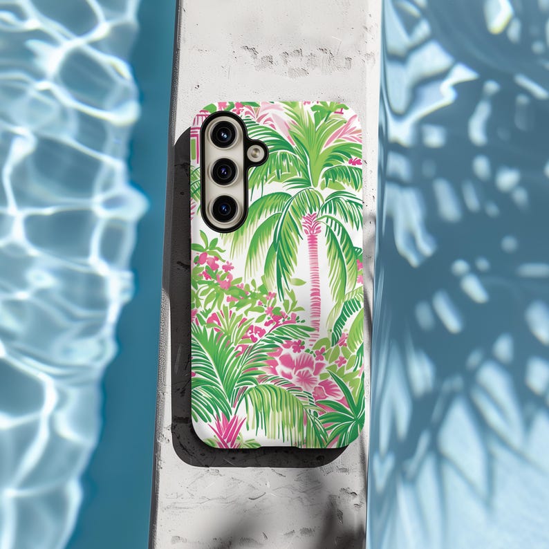 May include: A white phone case with a tropical print featuring pink flowers and green palm trees. The case has a cutout for a triple lens camera.