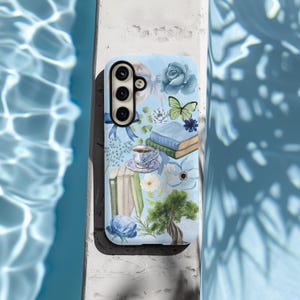 May include: A blue phone case with a watercolor design featuring flowers, books, a butterfly, and a teacup. The case has a camera cutout and a white background.