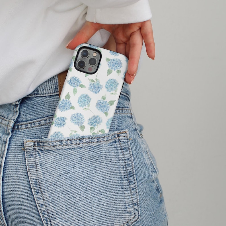 May include: A white phone case featuring a pattern of blue hydrangea flowers and green leaves. The case is partially visible in the back pocket of a pair of light blue denim jeans.