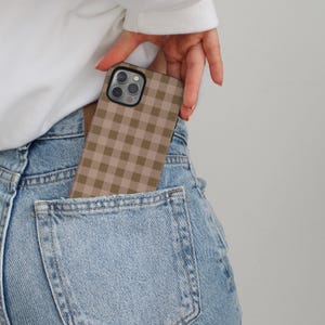 May include: A tan and brown gingham pattern phone case is partially visible in a denim jeans pocket. The phone case has a square pattern and a black border. The person's hand is holding the phone case.
