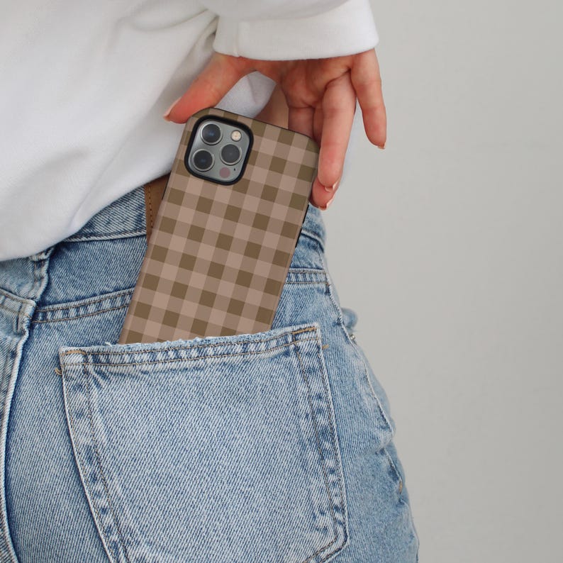 May include: A tan and brown gingham pattern phone case is partially visible in a denim jeans pocket. The phone case has a square pattern and a black border. The person's hand is holding the phone case.