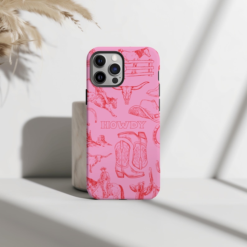 May include: Pink phone case with a red cowboy-themed design. The case features illustrations of a cowboy, boots, a hat, a bull skull, and the word "HOWDY". The case is designed for a smartphone.