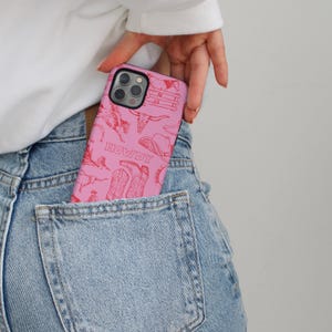 May include: Pink phone case with a red cowboy-themed design, including boots, a hat, and the word "HOWDY." The case is partially visible in a denim pocket.
