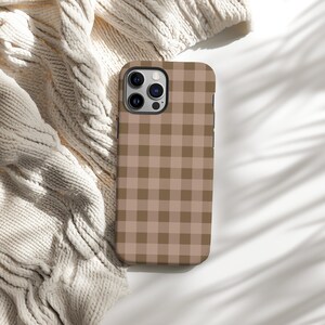 May include: A brown and tan checkered phone case. The case is made of a hard material and has a square pattern. The phone case is shown on a white surface with a cream-colored knitted sweater.