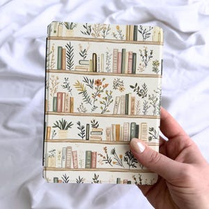 May include: A rectangular e-reader case featuring a repeating pattern of horizontal shelves filled with various books and botanical illustrations in shades of green, yellow, orange, and brown against a cream-colored background.