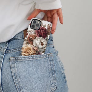 May include: A floral phone case featuring a design of peonies in shades of cream, burgundy, and peach. The case is in the pocket of a pair of light blue denim jeans.