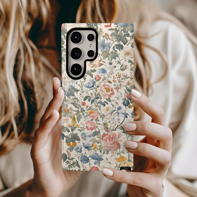 May include: A floral patterned phone case featuring a variety of flowers in shades of pink, blue, yellow, and green on a cream background. The case has a camera cutout and is held by a person.