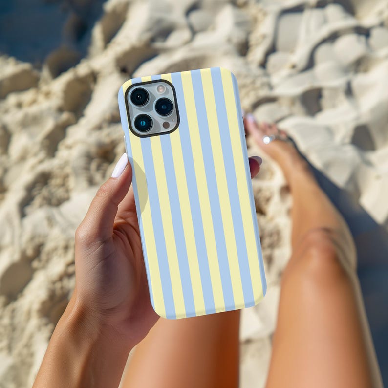Op de afbeelding: Een smartphonehoesje met een verticaal gestreept patroon in pastelgeel en lichtblauw. De hoes wordt in een hand gehouden, met zand op de achtergrond.