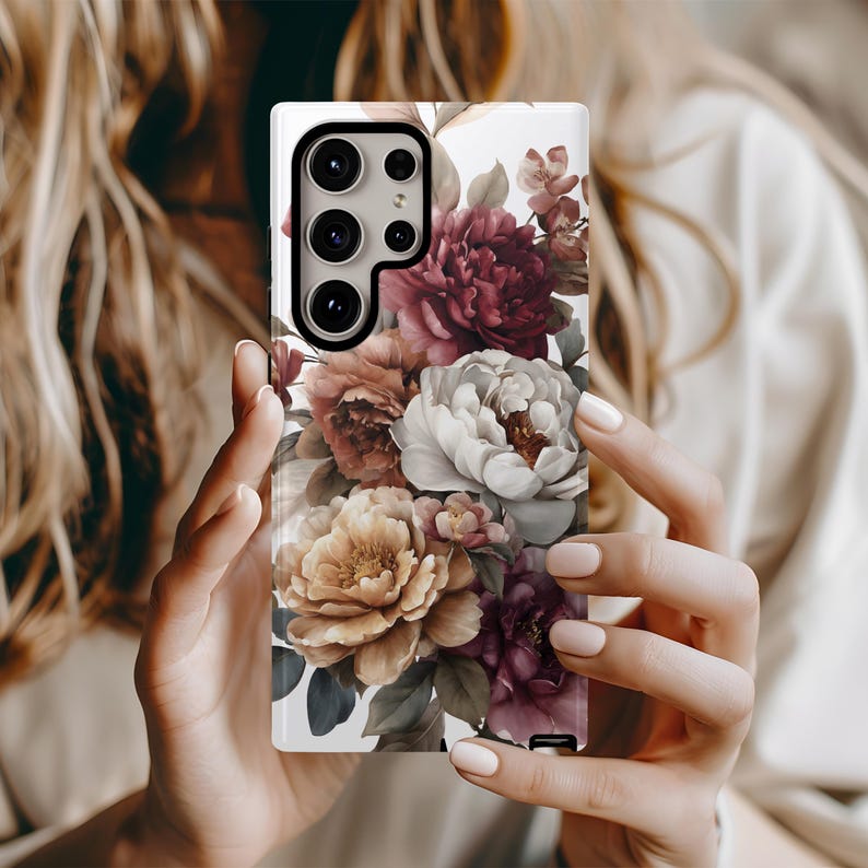 May include: A white phone case featuring a floral design with peonies in shades of burgundy, peach, and cream. The case has a glossy finish and a camera cutout. The floral arrangement is detailed and realistic, with green leaves.