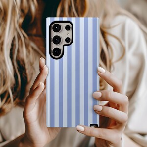 Peut inclure: Coque de t&eacute;l&eacute;phone &agrave; rayures verticales bleues claires et blanches. Le bo&icirc;tier est rectangulaire avec une ouverture pour l'appareil photo. La coque est tenue par une personne &agrave; la peau claire et aux longs cheveux blonds.