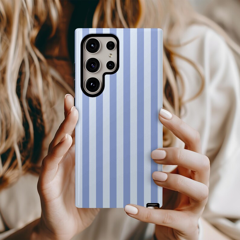Peut inclure: Coque de t&eacute;l&eacute;phone &agrave; rayures verticales bleues claires et blanches. Le bo&icirc;tier est rectangulaire avec une ouverture pour l'appareil photo. La coque est tenue par une personne &agrave; la peau claire et aux longs cheveux blonds.
