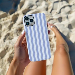 Peut inclure: Coque de smartphone &agrave; rayures verticales bleues claires et blanches. La coque est tenue dans une main, avec du sable de plage en arri&egrave;re-plan.