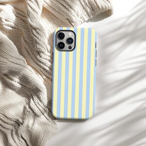 Op de afbeelding: Een smartphonehoesje met een verticaal streepjespatroon in pastelgeel en lichtblauw. De hoes heeft een zwarte cameramodule en zijknoppen. Het ligt op een cr&egrave;mekleurige gebreide stof.