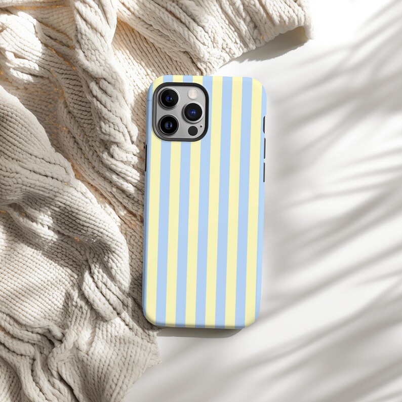 Op de afbeelding: Een smartphonehoesje met een verticaal streepjespatroon in pastelgeel en lichtblauw. De hoes heeft een zwarte cameramodule en zijknoppen. Het ligt op een cr&egrave;mekleurige gebreide stof.