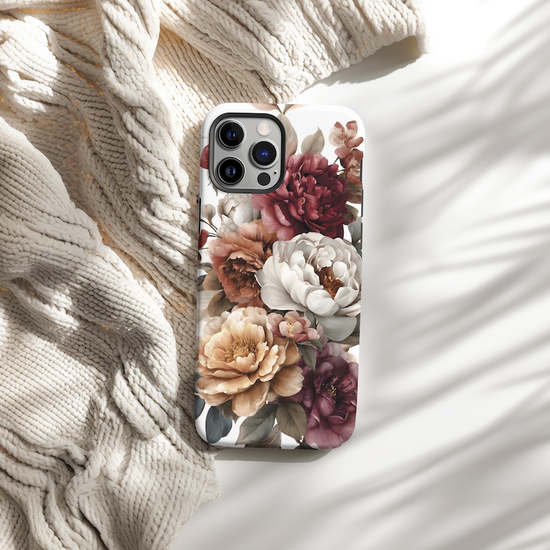 May include: A white phone case featuring a floral design with peonies in shades of burgundy, peach, cream, and yellow. The case has a glossy finish and a camera cutout. The design is a watercolor style.