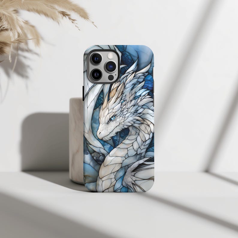 May include: A white and blue phone case featuring a dragon design. The dragon is white with blue accents and has a detailed, stained glass appearance.