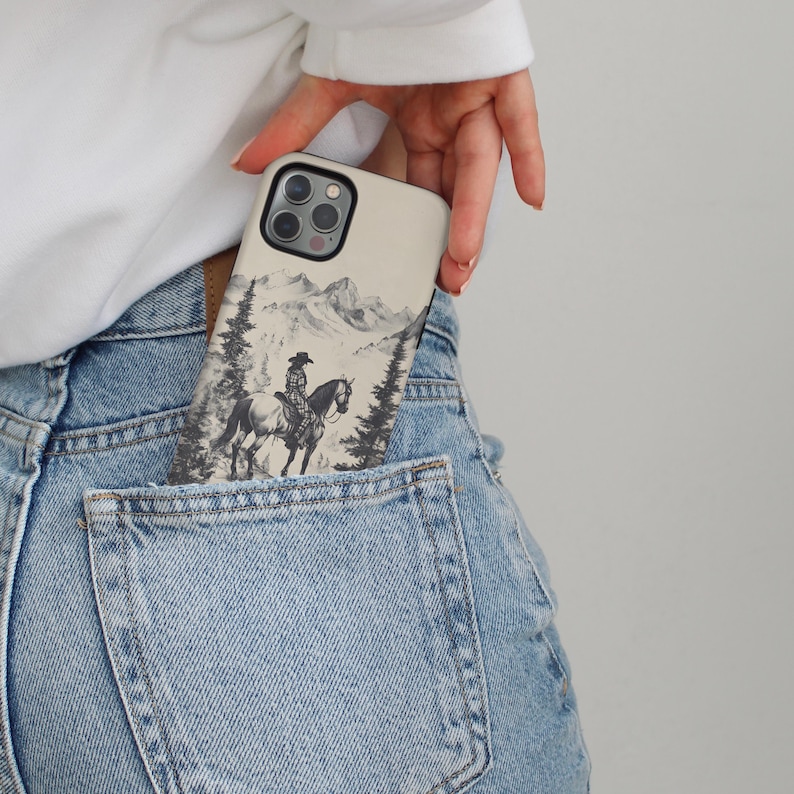 May include: A white phone case featuring a black and white illustration of a cowboy riding a horse through a snowy mountain landscape.