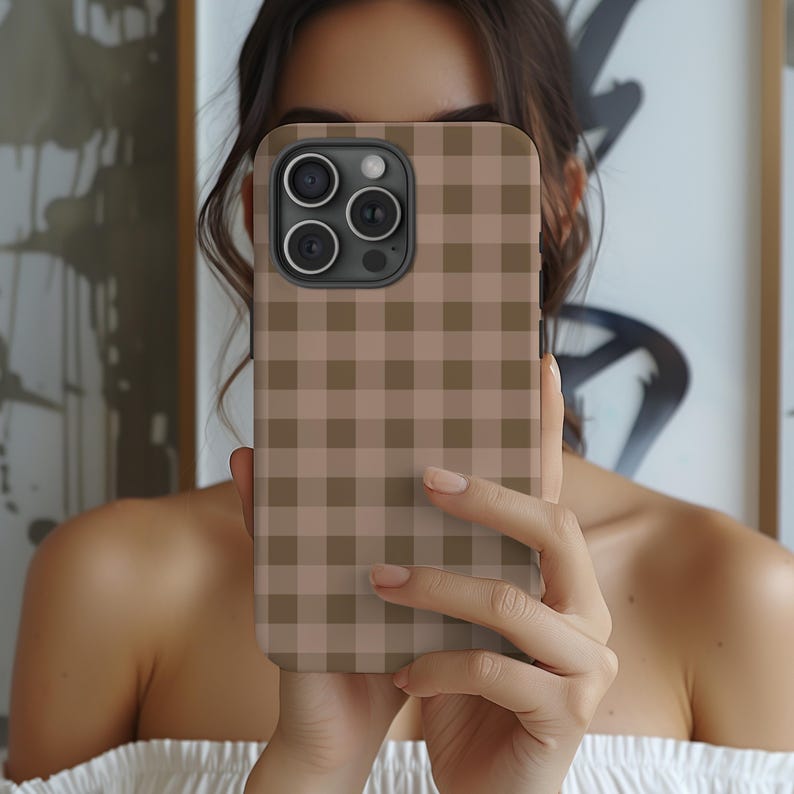 May include: A phone case with a brown and tan gingham pattern. The case has a camera cutout and is held in front of a person's face. The case is a neutral color and has a classic, timeless design.