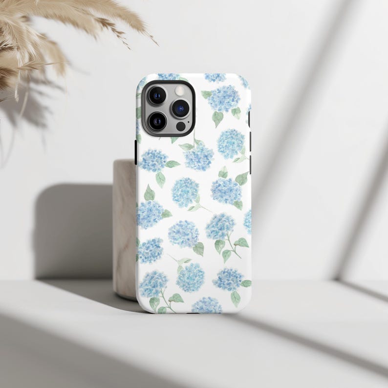 May include: A white phone case featuring a pattern of light blue hydrangea flowers with green leaves. The case has a black camera module and black side buttons. The phone case is standing upright on a white surface.