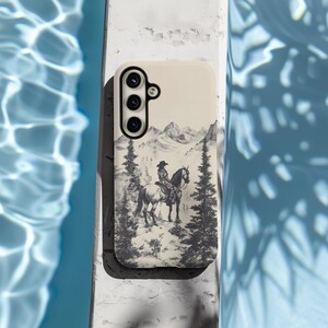 May include: A white phone case featuring a black and white illustration of a cowboy riding a horse in front of a mountain range.