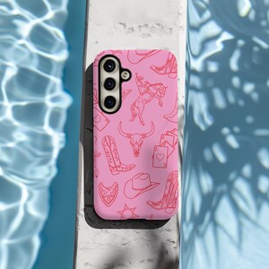 May include: Pink phone case with a pattern of cowboy boots, hats, horses, and playing cards. The case has a cutout for a triple camera lens.