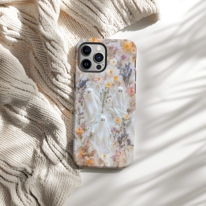 K&ouml;nnte beinhalten: Eine Handyh&uuml;lle mit floralem Design und wei&szlig;en Geistern. Die H&uuml;lle hat einen wei&szlig;en Hintergrund mit bunten Blumen und gr&uuml;nen Bl&auml;ttern. Die Geister sind wei&szlig; mit schwarzen Punktaugen. Die Handyh&uuml;lle liegt auf einer wei&szlig;en Oberfl&auml;che mit einer cremefarbenen Strickdecke.