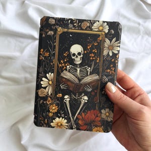 May include: A rectangular e-reader case featuring a dark, intricate illustration of a skeleton reading an open book. The skeleton is framed by a gold border and surrounded by a dense arrangement of white daisies, orange wildflowers, and red flowers.