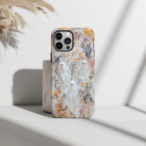 K&ouml;nnte beinhalten: Handyh&uuml;lle mit floralem Design und drei wei&szlig;en Geisterfiguren. Die H&uuml;lle hat einen wei&szlig;en Hintergrund mit gelben, rosa und lila Blumen. Die H&uuml;lle ist ein sch&uuml;tzendes Zubeh&ouml;r f&uuml;r ein Mobiltelefon.