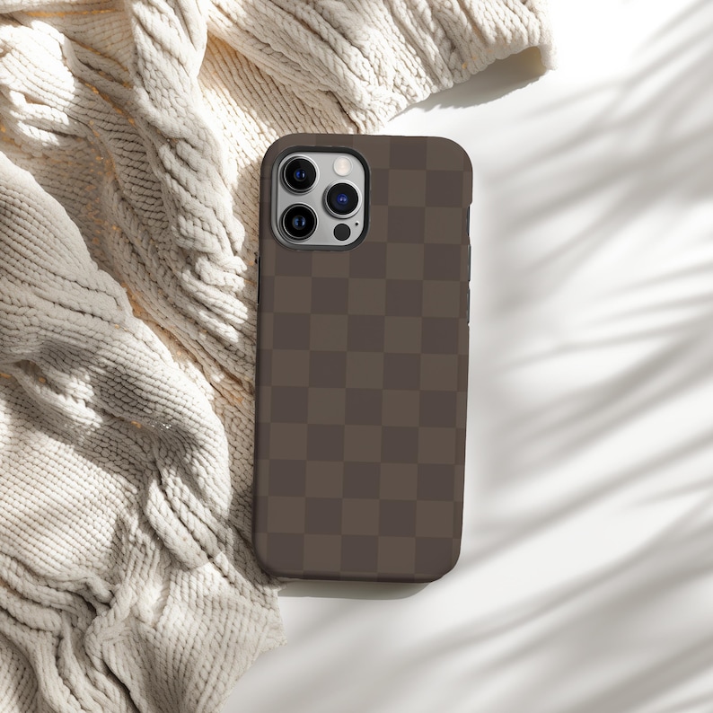 May include: A brown checkered phone case with a black camera bump and three lenses. The case is lying on a white surface with a white knitted blanket in the background.