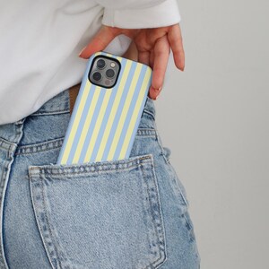 Op de afbeelding: Een telefoonhoesje met blauwe en gele verticale strepen is gedeeltelijk zichtbaar in een spijkerbroekzak. De telefoonhoes heeft een zwarte camerabehuizing. De persoon draagt een witte top met lange mouwen.