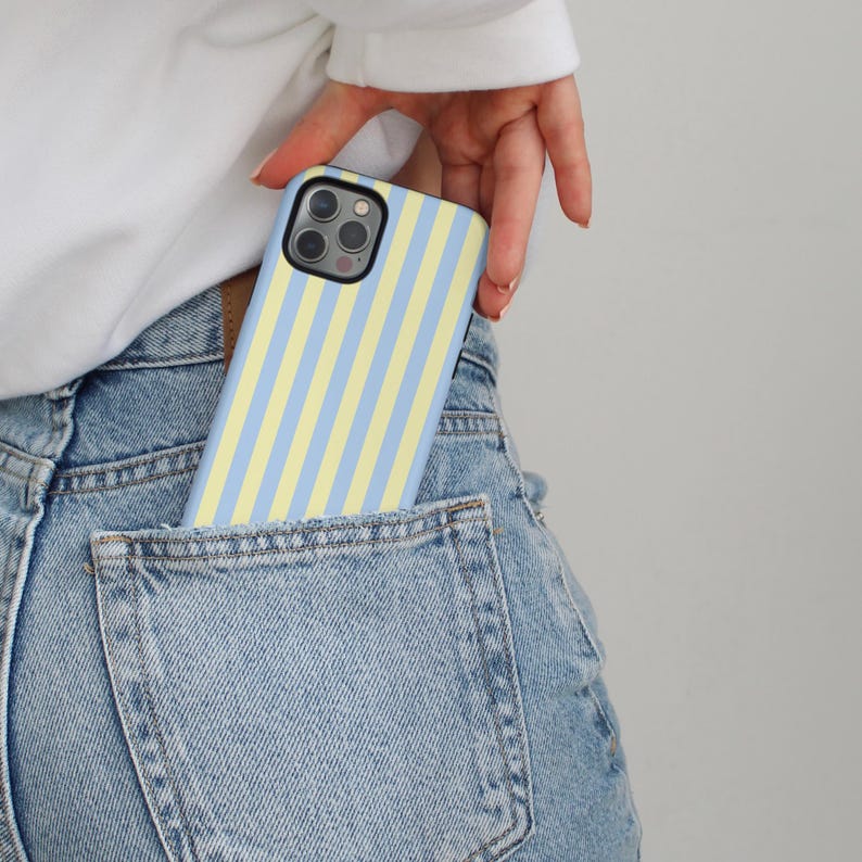 Op de afbeelding: Een telefoonhoesje met blauwe en gele verticale strepen is gedeeltelijk zichtbaar in een spijkerbroekzak. De telefoonhoes heeft een zwarte camerabehuizing. De persoon draagt een witte top met lange mouwen.