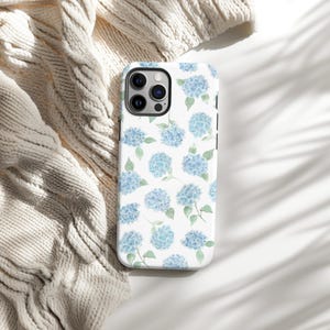 May include: A white phone case featuring a pattern of blue hydrangea flowers with green leaves. The case is shown on a white surface with a cream-colored knitted blanket in the background. The phone case has a glossy finish.