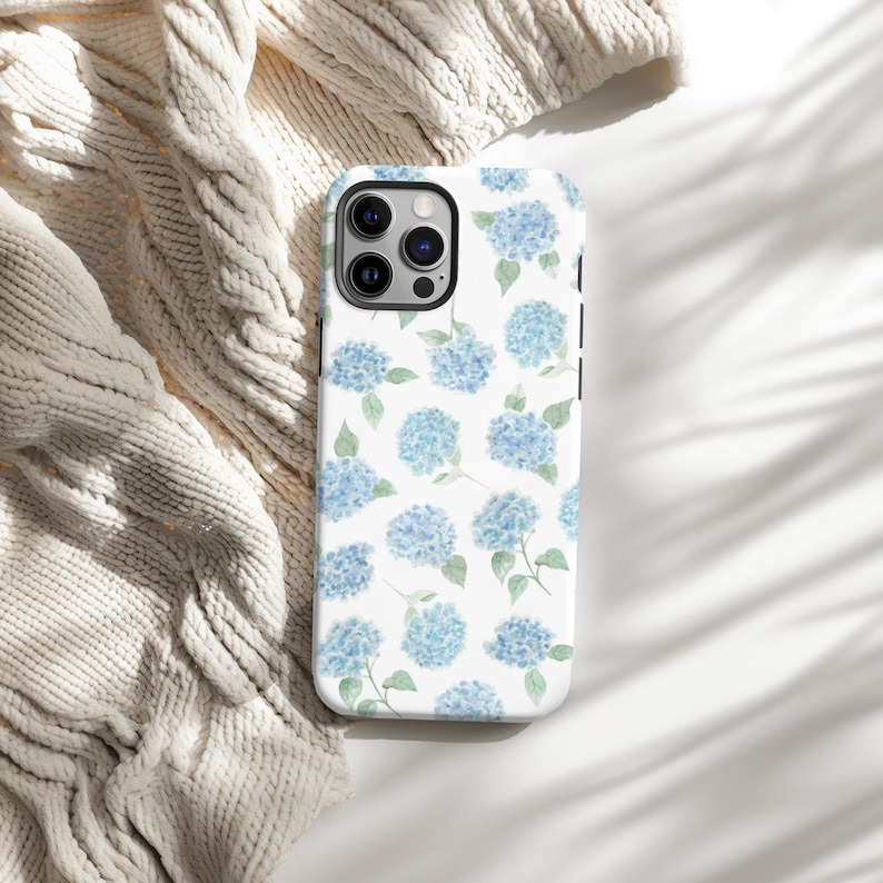 May include: A white phone case featuring a pattern of blue hydrangea flowers with green leaves. The case is shown on a white surface with a cream-colored knitted blanket in the background. The phone case has a glossy finish.