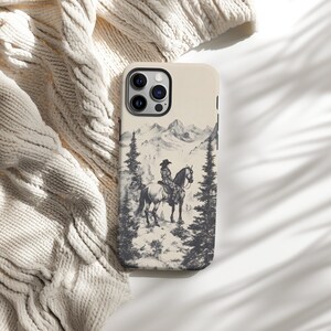 May include: A phone case featuring a black and white illustration of a person riding a horse in a mountainous landscape. The case has a cream background and a black camera lens.
