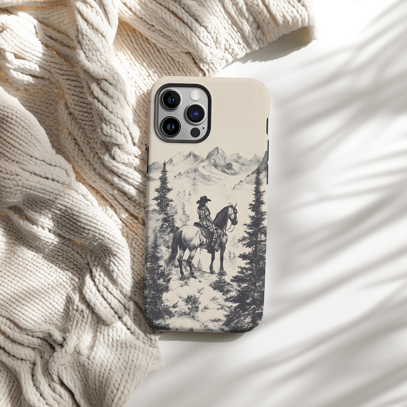 May include: A phone case featuring a black and white illustration of a person riding a horse in a mountainous landscape. The case has a cream background and a black camera lens.
