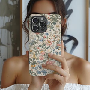 May include: A floral patterned phone case featuring a cream background with a repeating pattern of pink, blue, and yellow flowers and green leaves. The case has a camera cutout and is held up in front of a person.