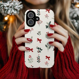 May include: A white phone case with a festive pattern of red bows, berries, and green sprigs. The case has a dual-lens camera cutout and is held by a person wearing a red sweater. The design evokes a holiday or winter theme.