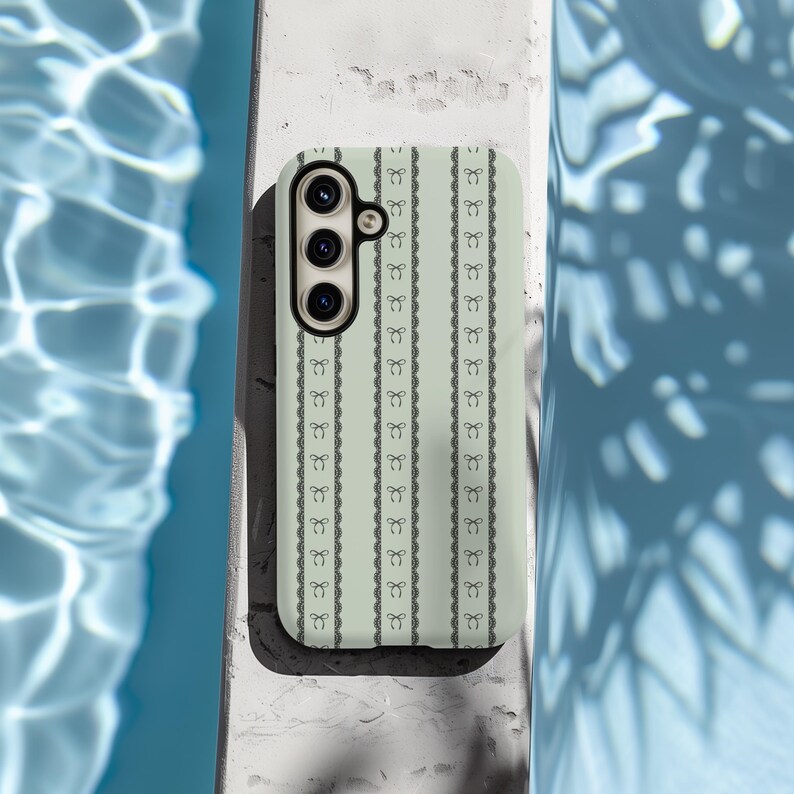 Pode incluir: Uma capa de telefone verde claro com um padr&atilde;o preto e branco de la&ccedil;os repetidos. A capa tem um recorte para a c&acirc;mera com tr&ecirc;s lentes.