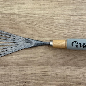 May include: A garden hand rake with a wooden handle and a gray grip that reads "Grace" with a small plant pot icon. The rake's tines are silver and spread out from a central point.