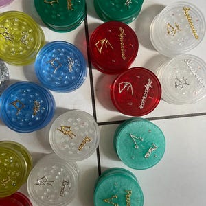 May include: A collection of 12 round, clear plastic containers with zodiac signs and symbols printed on them. Each container has a different color, including green, red, blue, yellow, and white. The containers are decorated with glitter and gold accents.