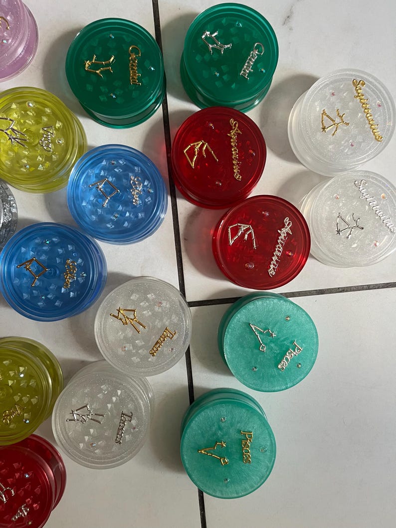 May include: A collection of 12 round, clear plastic containers with zodiac signs and symbols printed on them. Each container has a different color, including green, red, blue, yellow, and white. The containers are decorated with glitter and gold accents.