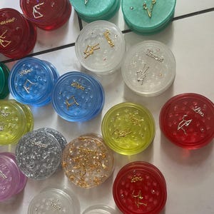 May include: A collection of 12 zodiac sign grinders. Each grinder is a different color and has a different zodiac sign on it. The signs are: Aquarius, Pisces, Taurus, Gemini, Cancer, Leo, Virgo, Libra, Scorpio, Sagittarius, Capricorn, and Aries.