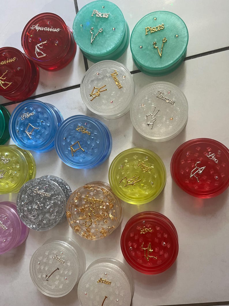 May include: A collection of 12 zodiac sign grinders. Each grinder is a different color and has a different zodiac sign on it. The signs are: Aquarius, Pisces, Taurus, Gemini, Cancer, Leo, Virgo, Libra, Scorpio, Sagittarius, Capricorn, and Aries.