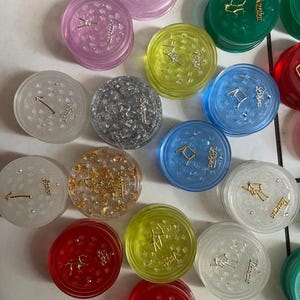 May include: Twelve round, clear plastic containers with zodiac symbols and glitter inside. Each container has a different zodiac symbol and color. The symbols are Gemini, Cancer, Leo, Virgo, Libra, Scorpio, Sagittarius, Capricorn, Aquarius, Pisces, Aries, and Taurus.