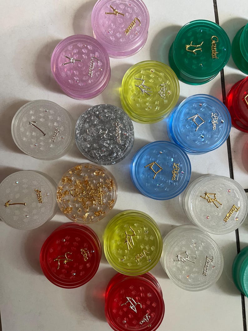 May include: Twelve round, clear plastic containers with zodiac symbols and glitter inside. Each container has a different zodiac symbol and color. The symbols are Gemini, Cancer, Leo, Virgo, Libra, Scorpio, Sagittarius, Capricorn, Aquarius, Pisces, Aries, and Taurus.