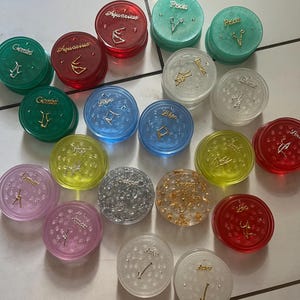 May include: A collection of 16 round, colorful, and clear grinder lids with zodiac signs and symbols. The lids are made of plastic and have a variety of colors, including red, green, blue, yellow, silver, and gold. Each lid has a different zodiac sign, including Gemini, Aquarius, Pisces, Libra, Virgo, and Sagittarius.