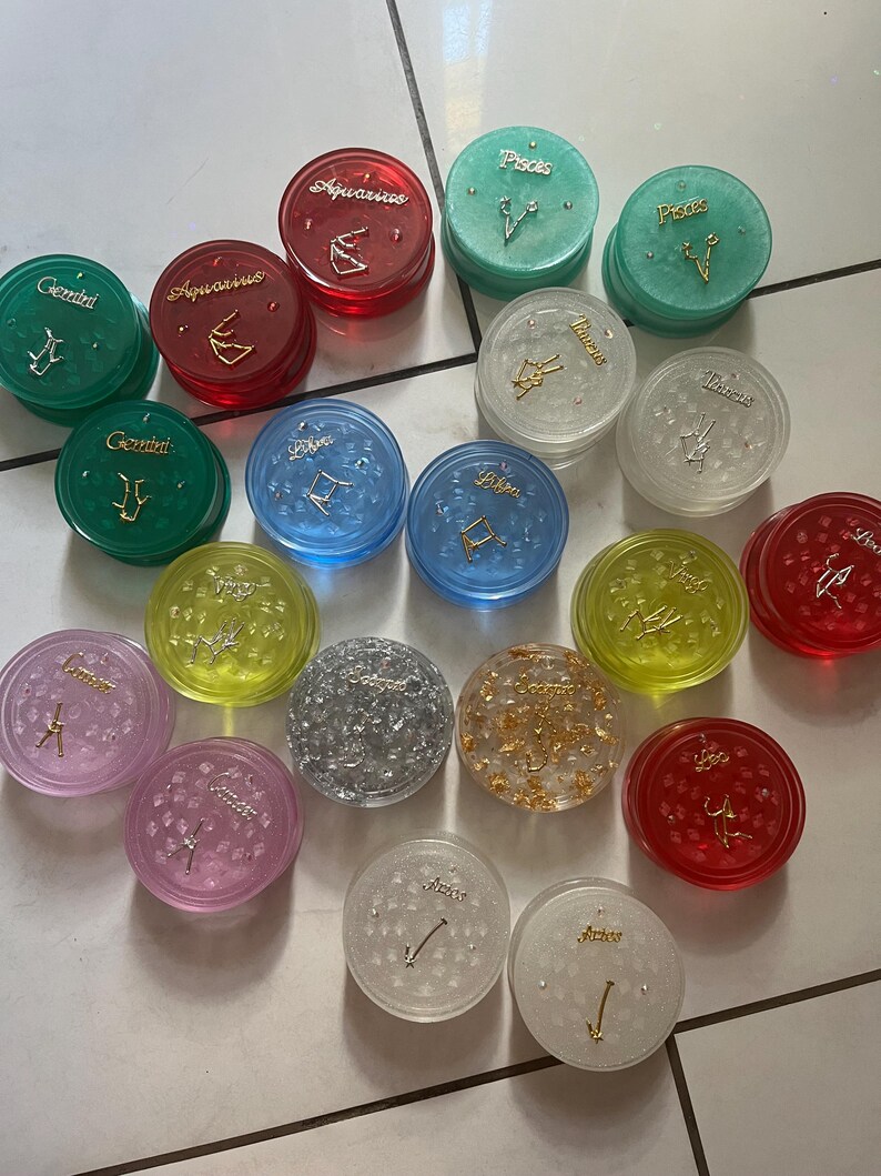May include: A collection of 16 round, colorful, and clear grinder lids with zodiac signs and symbols. The lids are made of plastic and have a variety of colors, including red, green, blue, yellow, silver, and gold. Each lid has a different zodiac sign, including Gemini, Aquarius, Pisces, Libra, Virgo, and Sagittarius.
