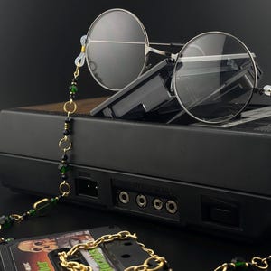 May include: Round silver-framed spectacles with a beaded chain featuring gold and green beads. The glasses rest on a black electronic device. A cassette tape with gold chain is in the foreground.