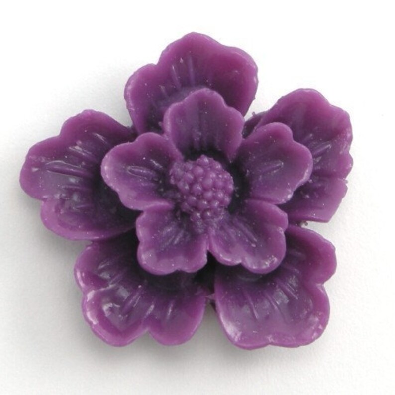 May include: A purple resin flower embellishment with a detailed, layered design. The flower has five petals and a center with a textured, raised design.