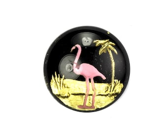 May include: A black button with a pink flamingo and a palm tree on a gold background.